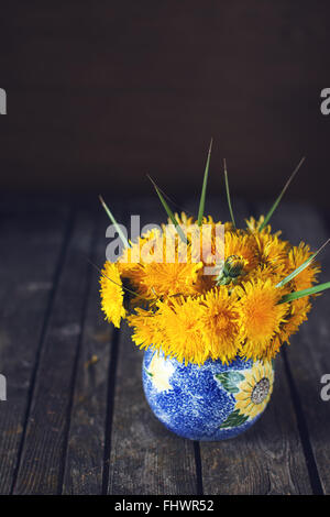 Helle, natürliche Löwenzahn Frühlingsstrauß auf einer dunklen hölzernen rustikale Oberfläche in blauer Vase drinnen Stockfoto