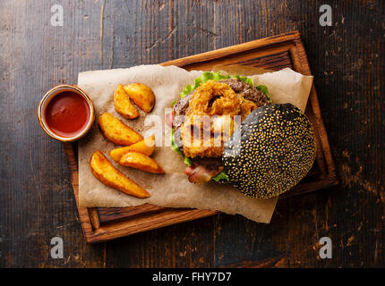 Schwarz-Burger mit Sesam Samen Brötchen Fleisch Speck Zwiebel Ringe, Pommes Frites und Kartoffelecken auf dunklem Holz Stockfoto