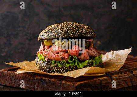 Roast Beef Burger Imbiss Snack auf Sesam Brötchen mit in Scheiben geschnittenen Pastrami und Gurke auf schwarzem Hintergrund Stockfoto