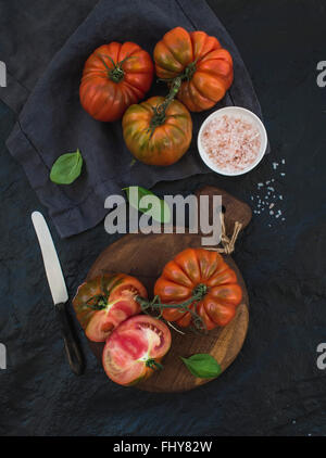 Frische reife Hairloom Tomaten und Basilikum Blätter auf rustikalem Holzbrett über schwarzem Stein. Ansicht von oben Stockfoto