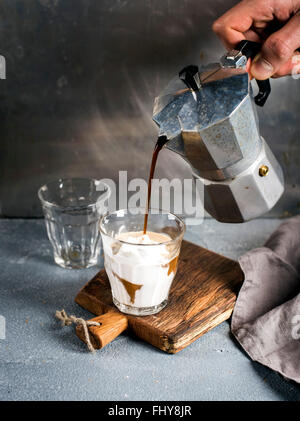 Ein Glas Kaffee mit Eis auf einem rustikalen Holzbrett. Getränk wird aus einer italienischen Moka-Stahltopf Besitz des Mannes Hand Gre gegossen. Stockfoto