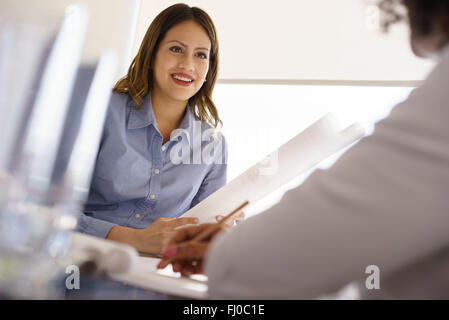 Team von zwei weiblichen Architekten, sitzen am Schreibtisch im Büro. Die Frauen sprechen und Lächeln, einen Bauplan zu überprüfen. Mittlere Aufnahme Stockfoto