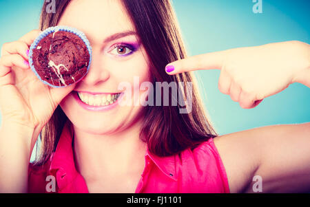 Diät süß Essen und Leute-Konzept. Lustige Frau hält Kuchen in der hand Spaß über ihr Auge mit Cupcake blauen Hintergrund Stockfoto