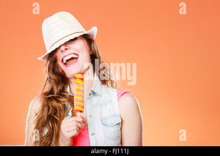 Sommer Urlaub Glück Konzept. Lustige fröhliche Frau, die Augen bedecken, mit Strohhut, essen Eis am Stiel Eis orange Hintergrund Stockfoto