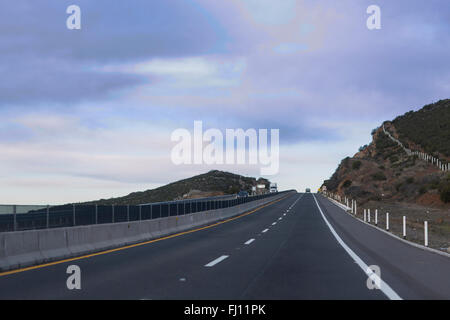 Unter dem kalten blauen Himmel erstreckt sich die Straße am Berg ohne Ende. Ein laufenden Autos macht die Ansicht lebendig. Stockfoto