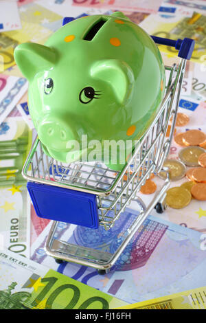 Grün-Sparschwein in einem blauen Einkaufswagen auf einem Hintergrund machte der Euro-Banknoten und-Münzen Stockfoto