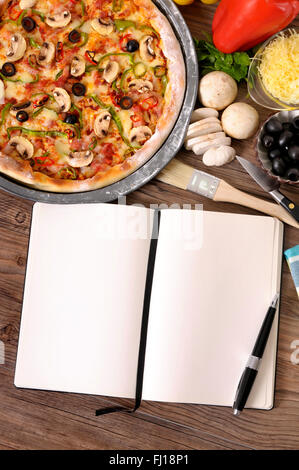 Frisch gebackene Pizza mit leeren Notebook oder Kochbuch umgeben von verschiedenen Zutaten. Stockfoto