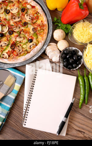 Frisch gebackene Pizza mit leeren Notebook oder Kochbuch umgeben von verschiedenen Zutaten. Stockfoto