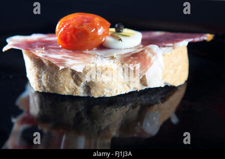 Spanische Tapa von Serrano-Schinken auf geschnittenem Brot mit Tomaten und gekochtem Ei serviert. Isoliert mit Reflexionen Stockfoto