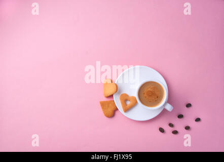 Tasse Kaffee auf rosa Papieroberfläche mit Sahped Herzkekse und ein paar Kaffeebohnen auf der Seite Stockfoto