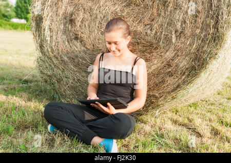 Schöne junge Mädchen mit Gerät sitzen auf dem Feld in der Nähe von Heuhaufen Stockfoto