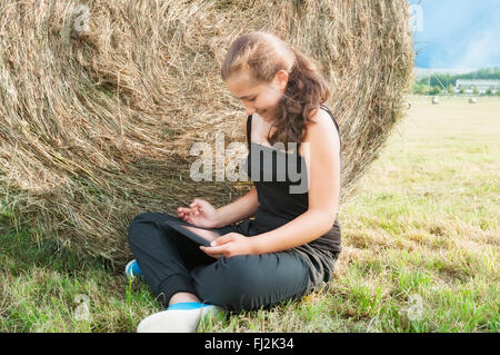 Mädchen mit Gerät sitzen auf dem Feld in der Nähe von Heuhaufen Stockfoto