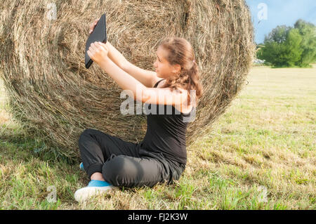 Junges Mädchen mit Gerät sitzen auf dem Feld in der Nähe von Heuhaufen mit Händen Stockfoto