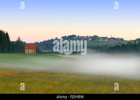 Wiesen im frühen Morgennebel, Schönberg Dorf im Hintergrund, Deutschland, Bayern, Oberbayern, Oberbayern, Schönberg Stockfoto