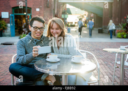 Zwei lächelnde junge Menschen suchen in Smartphone beim Sitzen an einem Tisch im Café im freien Stockfoto