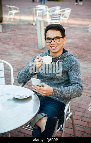 Junge asiatische Mann lächelnd in entspannenden outdoor Cafe Tasse Kaffee halten und messaging auf dem Handy Stockfoto