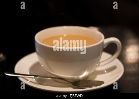 Eine Porzellantasse mit Kaffee und Sahne auf eine Untertasse mit einem Metalllöffel oben auf einem dunklen Tisch in einem Restaurant sitzen gefüllt Stockfoto