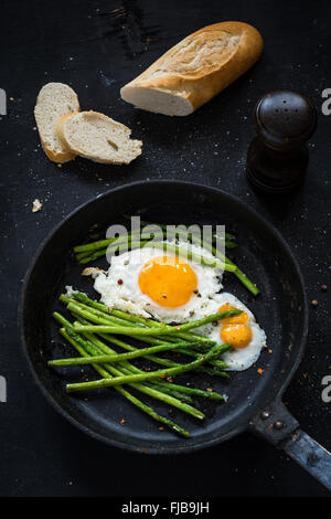 Grüner Spargel mit gebratenen Sonnenseite bis Ei in Pfanne und frischem Baguette auf schwarzem Hintergrund. Ansicht von oben, vertikale Zusammensetzung Stockfoto