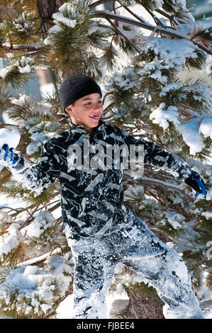 Niedliche kleine Junge spielt im Schnee versteckt vor einer Tanne. Stockfoto