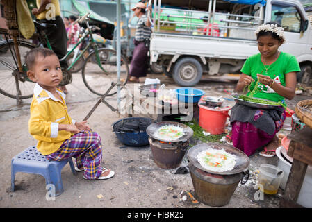 MANDALAY, Myanmar — Ein kleines Kind hilft ihrer Mutter, auf dem Fisch- und Blumenmarkt in Mandalay, Myanmar (Birma) Mahlzeiten zu kochen, die auf der Straße verkauft werden. Stockfoto