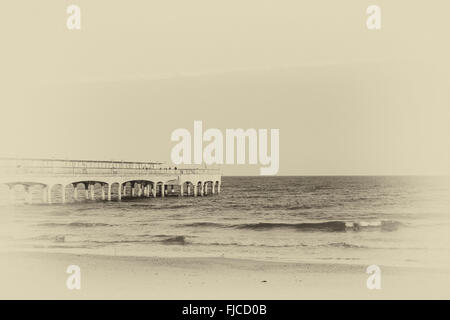 Ein schwarz-weiß Foto von einem Sonnenuntergang Licht, Blick auf einem Pier, aufgenommen am Strand mit Sand und keine Wolken am Himmel in Europa am Stockfoto