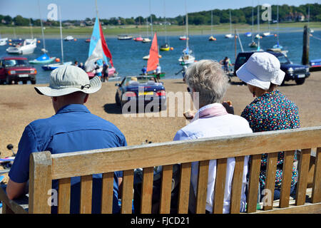 Drei ältere Menschen, die auf der Bank einen Happen zu essen haben und beobachten, wie das Boot vom Itschenor Slipway Itschenor, West Sussex, Großbritannien, gestartet wird Stockfoto