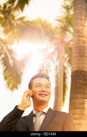 Ein Portrait eines glücklichen chinesischen Geschäftsmannes am Telefon zu sprechen. Stadtansicht im Hintergrund. Sonnenschein Leuchteffekt.