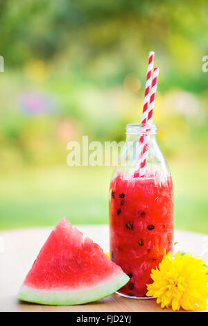 Erfrischende Sommer-Wassermelone trinken in einer Flasche mit dem Strohhalm trinken ein Stück Wassermelone und Blume auf Holztisch im Garten Stockfoto