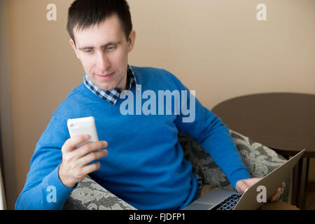 Porträt von lässig attraktive entspannt lächelnd junger Geschäftsmann mit Laptop und Handy in Sessel drinnen sitzen Stockfoto