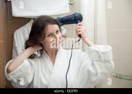 Porträt Jungen glücklich lächelnde Frau mit weißen Bademantel mit Haartrockner im Badezimmer, immer bereit, trocknen Sie ihr Haar Stockfoto