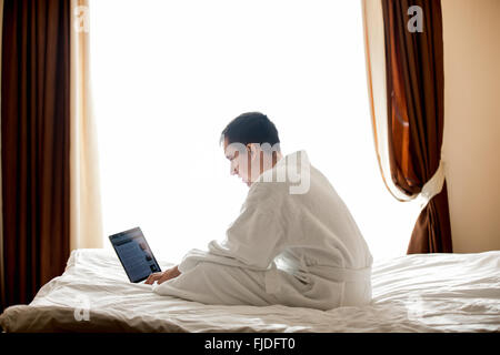 Porträt der attraktive junge Mann sitzt auf Bett mit weißen Bademantel, konzentrierte sich auf die Arbeit, männliches Model mit Laptop im hotel Stockfoto