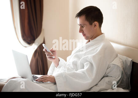 Porträt von attraktiven entspannten Jüngling tragen weiße Bademantel sitzen auf dem Bett mit Laptop und Handy, männliches model Stockfoto