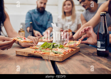 Schuss von Pizza am Tisch, mit einer Gruppe junger Leute herumsitzen und hob einen Teil hautnah. Freunden feiern und Essen Stockfoto