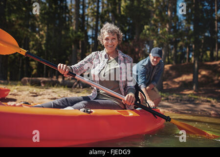 Porträt der glückliche ältere Frau in einem Kajak Paddel halten. Frau mit Mann im Hintergrund am See Kanu. Stockfoto