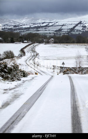 Spuren entlang der Länge der ländlichen schneebedeckte Fahrbahn in Flintshire mit der Clwydian Hügel oder Clwydian Hügel Hügel und Moel Famau in der Ferne, Flintshire, Wales, Großbritannien Stockfoto