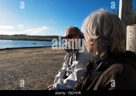 Ehepaar im Ruhestand sitzen auf einer Bank am Meer am Rock, Cornwall, UK. Stockfoto