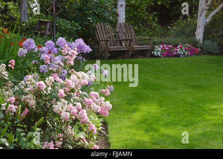 Cluster von rosa und weißen Tee Rosen von einer saftig grünen Wiese mit zwei rustikale Stühle warten auf Sie im Hintergrund. Stockfoto