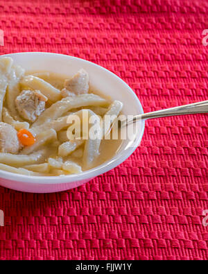Eine weiße Schüssel voller hausgemachter Hähnchen und Nudeln auf eine rote Platzdeckchen. Stockfoto