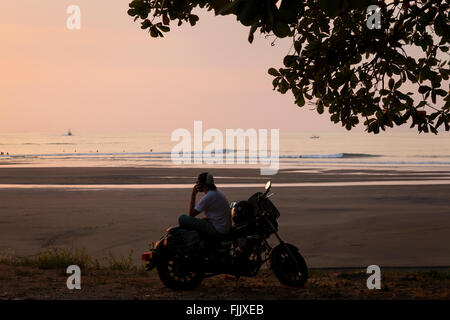 Ein Mann sitzt auf einem Motorrad vor dem Strand bei Sonnenuntergang in Quepos, Provinz Puntarenas, Costa Rica. Stockfoto