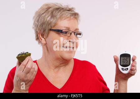 Glücklich lächelnd Seniorin hält Blutzuckermessgerät mit positiven Ergebnis der Messung Zucker Ebene und frischen Kuchen, Stockfoto