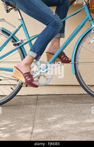 Woman's Beine treten ein Fahrrad auf einem Bürgersteig. Sie trägt Jeans und Keil - Sandaletten. Gesunder Lebensstil spaß Sommermode in der Stadt. Stockfoto