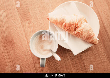 Cappuccino mit Croissant. Tasse Kaffee steht auf Holztisch in der Cafeteria, Nahaufnahme Foto mit Tiefenschärfe, Draufsicht, retr Stockfoto