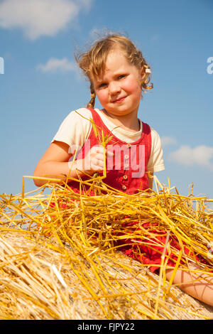 Kleines Mädchen sitzt auf einem Strohballen bei bewölktem Himmel blau, Deutschland Stockfoto