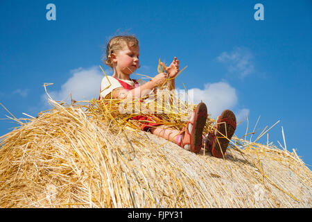 Kleines Mädchen sitzt auf einem Strohballen bei bewölktem Himmel blau, Deutschland Stockfoto