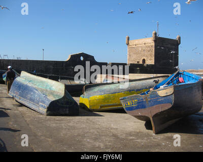 Blau und Gelb Fischerboote liegen auf ihren Seiten in Essaouira in Marokko mit den alten portugiesischen Festung im Hintergrund. Stockfoto