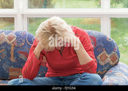 Verzweifelte blonde Frau mittleren Alters sitzt auf dem Sofa mit gesenktem Kopf. Stockfoto