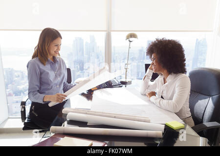 Team von zwei weiblichen Architekten, sitzen am Schreibtisch im Büro. Die Frauen sprechen und Lächeln, einen Bauplan zu überprüfen. Mittlere Aufnahme Stockfoto