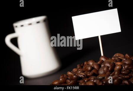 Ein Haufen von Kaffeebohnen mit einem leeren Zeichen auf sie und eine weiße Kaffeetasse im Hintergrund Stockfoto