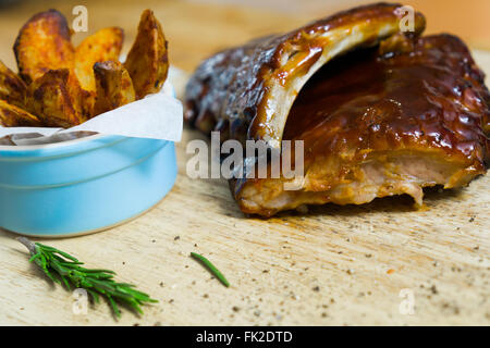 Nahaufnahme zeigt zwei Grill Schweinerippchen, glasiert mit rauchiger Sauce und Kartoffel Wedges auf einem Holztisch Stockfoto