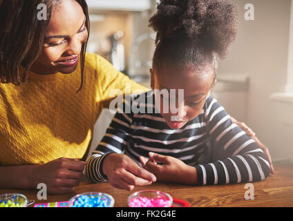 Glückliche Mutter in gelben Bluse und Kind arbeiten mit Perlen in verschiedenen Farben in Gläsern auf Holztisch Stockfoto